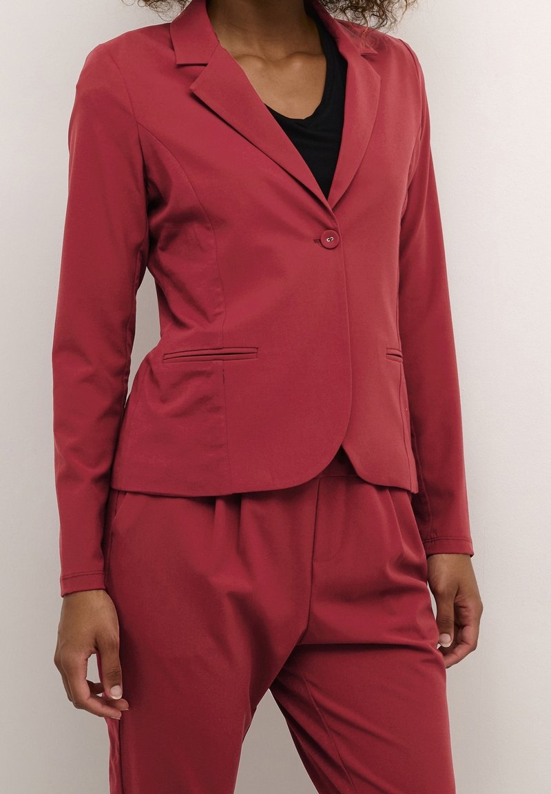 Blazer rouge avec une silhouette ajustée, fermeture à un bouton et deux poches avant. Fabriqué en tissu lisse avec une finition mate.