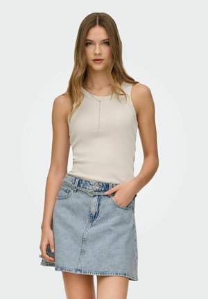 Giovane donna con capelli castano chiaro lunghi, indossa un top beige senza maniche e una gonna di jeans azzurri, in piedi con una mano nella tasca.