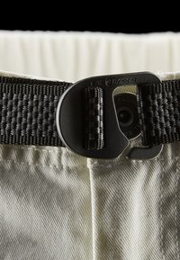 Weiße Hosen zeichnen sich durch einen strukturierten Stoff aus und haben einen schwarzen gewebten Gürtel. Der Gürtel verfügt über einen metallischen Verschluss mit einem kreisförmigen Design und einem Branding-Detail.