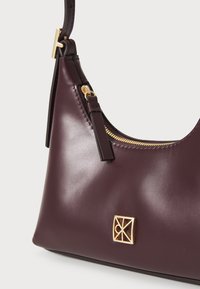 Geantă de mână din piele bordo cu un fermoar și accesorii din aur, având un plăcuță rectangulară cu logo și o textură netedă, lucioasă.