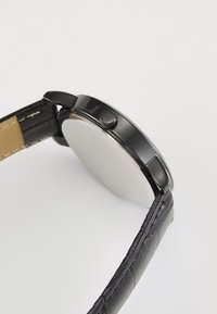 Black watch with a round case, stainless steel back, and textured black leather strap. Includes a side crown for adjustments.