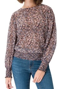 Femme portant un chemisier à manches longues à motifs floraux avec des poignets et un ourlet smockés, associé à un jean en denim bleu.