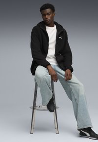 Schwarze Fleecejacke mit Kapuze, weißes T-Shirt, hellblaue Jeans und schwarze Sneakers. Model sitzt auf einem verchromten Barhocker vor einem neutralen Hintergrund.