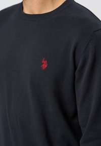 Marinblå stickad tröja med rund halsringning och ribbade mudd. Har en liten röd broderad logotyp på vänster bröst. Mjuk textur, avslappnad passform.