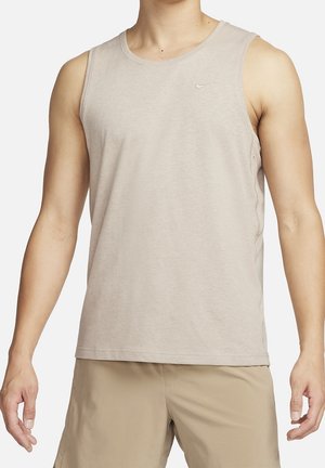 Mand iført lys beige ærmeløs Nike tanktop og khaki shorts, stående med arme afslappet ned langs siderne mod hvid baggrund.