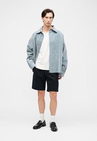 Giovane uomo che indossa una giacca oversize azzurro chiaro, t-shirt bianca, pantaloncini neri, calzini bianchi e mocassini neri, in piedi davanti a uno sfondo bianco.
