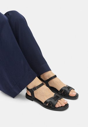 Black leather sandals featuring a strap around the ankle, cut-out design on the upper, and a flat sole, paired with navy pants.
