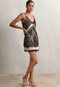 Rochie slip din dantelă în negru și roz pal cu bretele ajustabile, tiv ondulat și suprapunere din dantelă florală. Modelul pozează pe un fundal neutru.