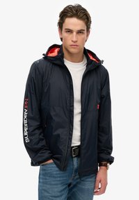 Chaqueta cortavientos azul marino con capucha forrada en naranja, cierre frontal de cremallera, detalle del logo en la manga y puños elásticos; combinada con una camisa blanca y jeans.