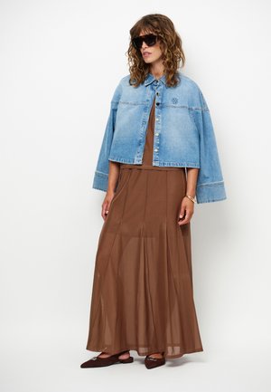 Vrouw draagt een oversized lichtblauwe spijkerjack, een bruin maxi-jurk, grote zwarte zonnebril en bruine puntige platte schoenen, staand tegen een witte achtergrond.