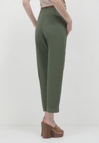 Persona che indossa pantaloni a vita alta di colore verde oliva con orli risvoltati e mule con tacco a piattaforma marroni con borchie dorate, in piedi su un pavimento bianco.