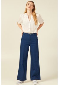 Camicia bianca con colletto aperto e maniche corte a palloncino, pantaloni blu a gamba larga e sneakers bianche su uno sfondo neutro.