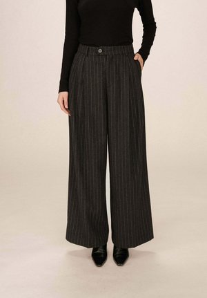 Femme portant un pantalon large en laine à rayures grises foncées taille haute, des chaussures noires à bouts pointus et un haut noir à manches longues, se tenant avec une main dans la poche.