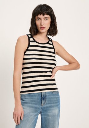 Frau mit schulterlangem dunklem Haar, die ein ärmelloses, schwarz-weiß gestreiftes Oberteil und blaue Jeans trägt, mit Hand in der Hüfte stehend.