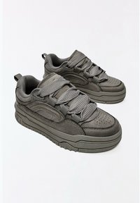 Paire de baskets épaisses en daim gris avec des lacets épais texturés et des semelles en caoutchouc à motifs sur fond blanc uni.