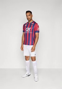 Φανέλα ποδοσφαίρου με κόκκινες και μπλε ρίγες και κοντά μανίκια, λευκό σορτς και λευκές κάλτσες. Μαύρα και λευκά παπούτσια ποδοσφαίρου με υφή λεπτομέρειας.