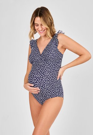 Smiling pregnant woman in navy blue floral maternity swimsuit with ruffled straps, holding belly with one hand and resting other on back.