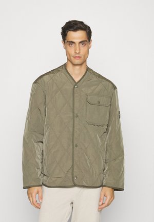 Άνδρας που φοράει πετσετωμένο ελαιόχρωμο μπουφάν με κουμπιά και τσέπη στο στήθος, φορώντας μπεζ παντελόνι, στέκεται μπροστά σε λευκό φόντο.