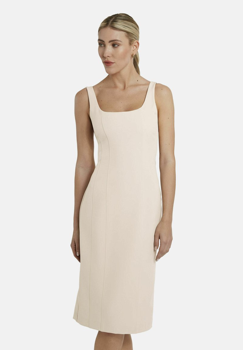 Vrouw met blond haar in een lage paardenstaart draagt een mouwloze, nauwsluitende, knie-lange beige jurk met een vierkante halslijn, staand tegen een witte achtergrond.