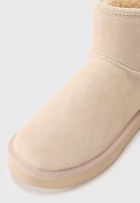 Beige semišové kotníkové boty s kulatou špičkou, měkkou fleecovou podšívkou a prošívanými detaily na límci a podrážce. Texturovaný povrch.