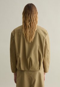 Chaqueta de color caqui con un corte relajado y mangas largas, hecha de una tela suave. Presenta un dorso fruncido para añadir forma.