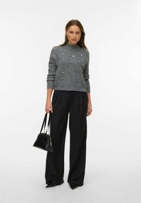 Grijze gebreide trui met een hoge halslijn, versierd met zilveren bloemmotieven, gecombineerd met zwarte wijde broekspijpen en een kleine zwarte handtas.