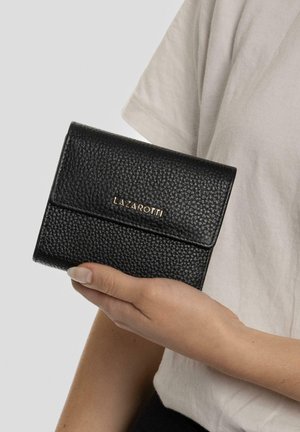 Cartera de piel negra con un acabado texturizado, que presenta una solapa frontal y un logotipo dorado en relieve. Sostenida en una mano frente a un fondo neutro.