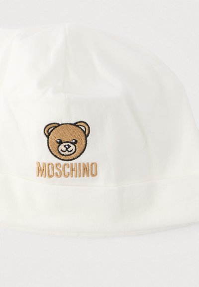 Λευκό βαμβακερό σκουφάκι με κεντημένο πρόσωπο αρκούδας και το κείμενο "Moschino" σε χρυσό κάτω από την αρκούδα, με απαλή υφή και στρογγυλεμένο σχήμα.