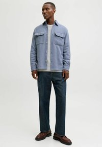 Lys blå skjorte med knapper og to brystlommer, brukt over en grå topp. Mørkeblå jeans med rette ben og brune snørebåndsko fullfører antrekket.