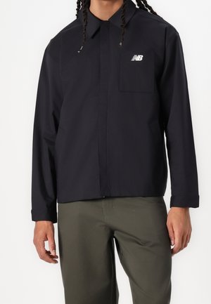 Persona che indossa una giacca nera New Balance con cerniera e tasca sul petto e pantaloni verde oliva su uno sfondo bianco neutro.