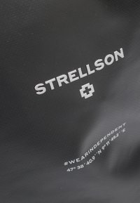 Bolso negro texturizado con el logo en relieve en blanco "STRELLSON" y símbolo de cruz. Presenta un hashtag y coordenadas impresas en blanco en la parte inferior.