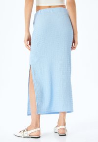 Lichtblauwe, gestructureerde maxi-jurk met een hoge split aan de rechterkant, gecombineerd met witte sandalen met een metallic accent.