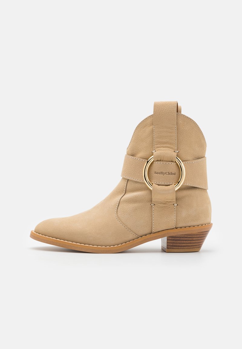 Beige ankle boot with a rounded toe, made of suede. Features a wide strap with a gold circular accent and a low wooden heel.