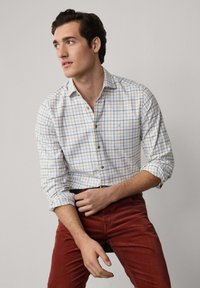 Camicia bianca con un motivo a griglia blu e arancione, maniche arrotolate, abbinata a pantaloni in velluto a coste color ruggine, cintura nera semplice.