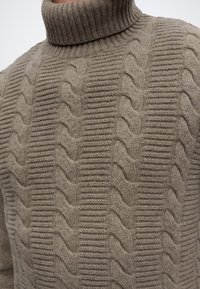 Pull à col roulé beige tricoté avec des motifs torsadés verticaux et un col côtelé.