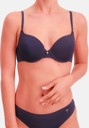 Mujer con un conjunto a juego de sujetador y ropa interior azul oscuro, con una mano apoyada en el estómago sobre un fondo liso.