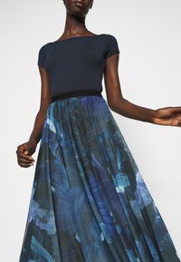 Kvinna klädd i en figurnära marinblå kortärmad topp och en svävande plisserad kjol med ett blått blommönster.