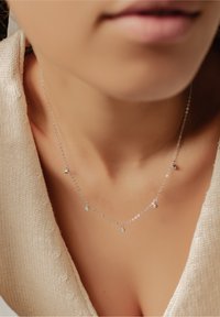 Srebrny naszyjnik z delikatnym łańcuszkiem, z małymi, okrągłymi zawieszkami ozdobionymi błyszczącymi akcentami. Noszony blisko dekoltu.
