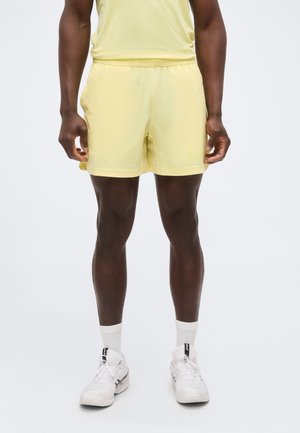 Uomo con pantaloncini e maglietta sportivi giallo chiaro, calzini bianchi e scarpe da corsa bianche, in piedi su sfondo bianco.