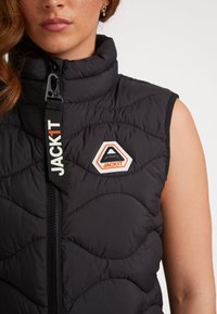 Svart pufferväst med ett quiltat mönster, hög krage och framdragkedja. Har en logotyp patch och kontrasterande dragkedjehandtag.