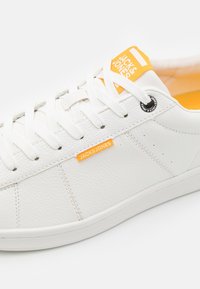 Weiße Leder Sneaker mit flachen Schnürsenkeln, orangefarbener Markenaufschrift und strukturiertem Obermaterial. Runde Ösen und leichte Gummisohle.