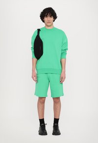 Un outfit format dintr-un hanorac mint green și pantaloni scurți, asortat cu o geantă neagră tip crossbody, având o croială lejeră, textură netedă și un design minimalist.