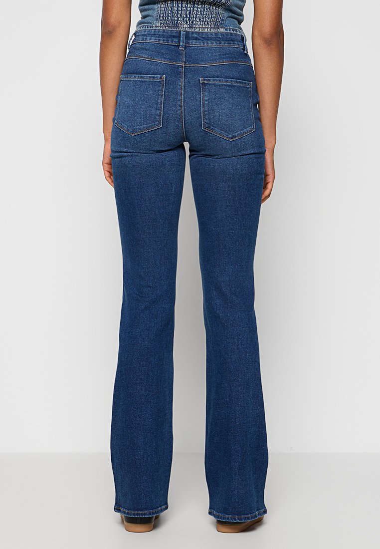 Femme portant un jean en denim bleu foncé taille haute avec des poches arrière, se tenant face à un mur blanc avec des pieds visibles dans des chaussures marron.