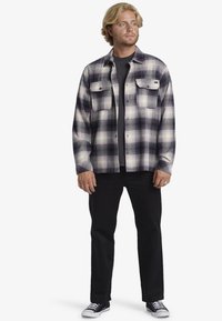 Zwart en paars geruiten flanellen shirt met twee borstzakken, gelaagd over een zwart shirt, gecombineerd met zwarte broek en zwarte sneakers.