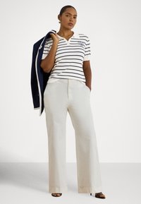 Sieviete valkā baltu un tumši zilu svītrainu polo kreklu, krēmkrāsas plaša piegriezuma bikses un melnas augstpapēžu kurpes, pār plecu nēsā tumši zilu žaketi.