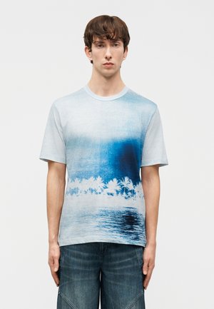 Νέος άνδρας που φοράει μπλε και λευκό κοντομάνικο μπλουζάκι με εκτύπωση φοίνικα και νερού, συνδυασμένο με σκούρα τζιν σορτς.