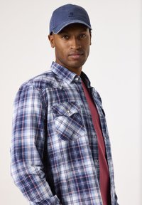 Camisa de cuadros azules con bolsillos abotonados sobre una camiseta burdeos, acompañada de una gorra azul marino. Los materiales parecen basados en algodón.