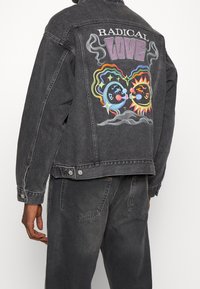 Svart denimjacka med flerfärgad broderad design av sol och måne på baksidan med texten "RADIKAL KÄRLEK" ovanför.