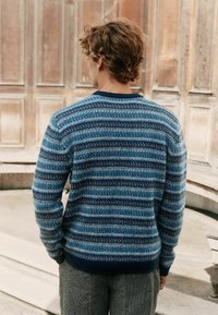 Junger Mann mit welligem braunem Haar, der einen blau gestreiften Strickpullover und graue Hosen trägt, steht vor einer Holzverkleidung.