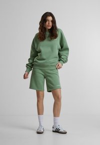 Hele roheline oversized sviiter ribatud mansettide ja äärega, koos sobivate lühikeste püksidega. Vasakul küljel valge logo detail; mudel kannab spordijalatseid.